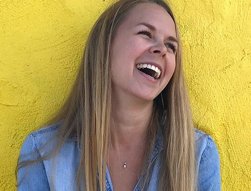 Girl with dark blonde hair laughing, up against a bright yellow wall and wearing denim shirt with solitaire diamond fine necklace