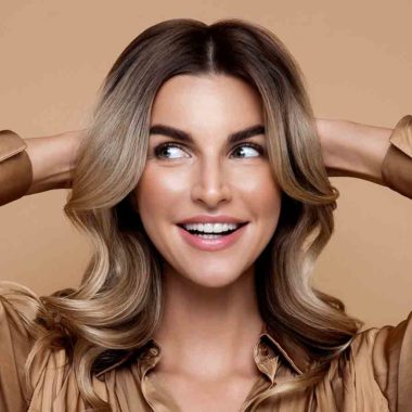 Creamy Blonde_image 1 Female with creamy blonde, wavy hair, wearing a bronze blouse, holding both hands behind head.