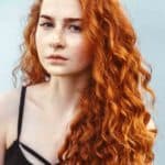 Long, curly, red head female with freckles, standing against white wall